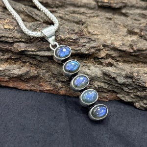 May include: A silver pendant necklace featuring five oval-shaped labradorite stones. The stones display iridescent blue and green hues. The pendant is suspended from a silver chain. The background is a textured, dark brown surface.