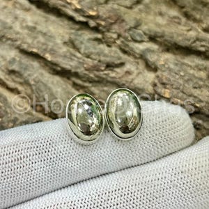 Puede incluir: Un par de pendientes de botón ovalados plateados. Los pendientes tienen una superficie pulida y reflectante. Los pendientes se muestran sobre una superficie de tela blanca, con un fondo de madera borroso.