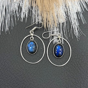 Puede incluir: Pendientes de aro plateados con piedras preciosas ovaladas de color azul oscuro. Las piedras tienen una cualidad iridiscente y brillante. Los pendientes se muestran sobre una superficie negra, con hebras blancas y marrones claras de fondo.