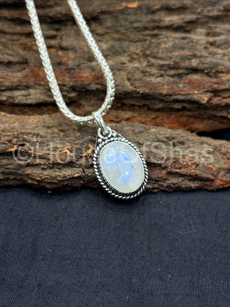 May include: A silver necklace with a teardrop-shaped pendant. The pendant features a large, oval, iridescent stone set in a silver bezel with a twisted rope design. The chain is a silver snake chain.