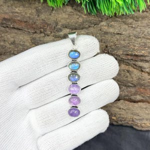 May include: A silver pendant featuring six oval gemstones in a vertical row. The gemstones transition in color from blue to purple and lavender. The pendant is displayed against a natural wood background.