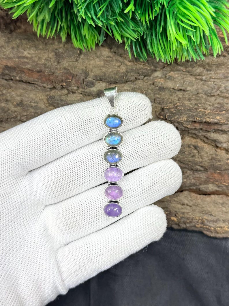 May include: A silver pendant featuring six oval gemstones in a vertical row. The gemstones transition in color from blue to purple and lavender. The pendant is displayed against a natural wood background.