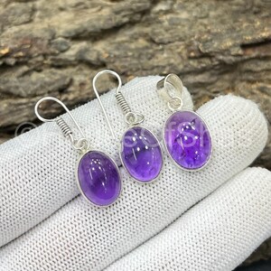 May include: A jewelry set featuring a pair of amethyst earrings and a matching pendant. Each piece has a silver setting and a polished, oval-shaped purple gemstone. The earrings have a hook design, while the pendant has a loop.