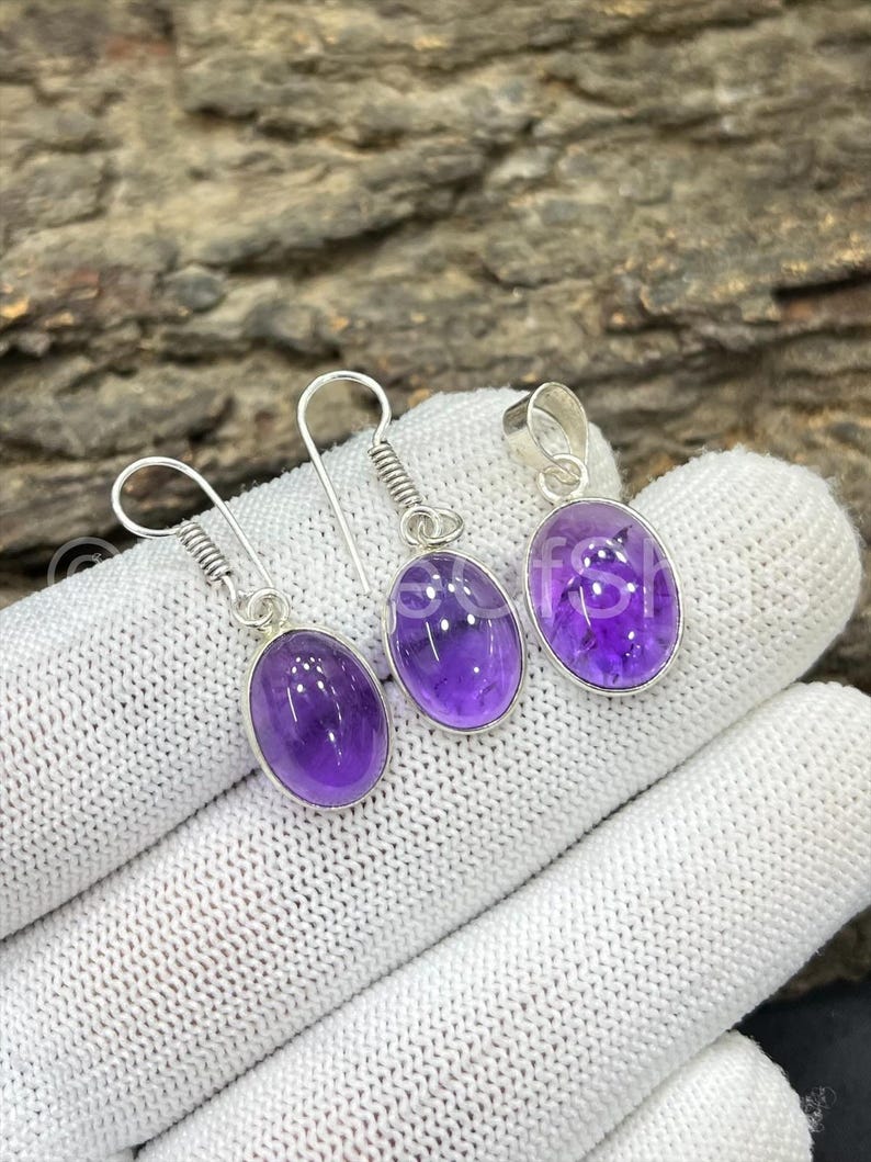 May include: A jewelry set featuring a pair of amethyst earrings and a matching pendant. Each piece has a silver setting and a polished, oval-shaped purple gemstone. The earrings have a hook design, while the pendant has a loop.
