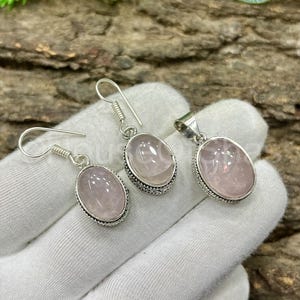 May include: A jewelry set featuring a pair of earrings and a pendant. Each piece has a light pink, oval-shaped stone set in a silver-colored metal frame. The earrings have a hook design, and the pendant has a loop for a chain.
