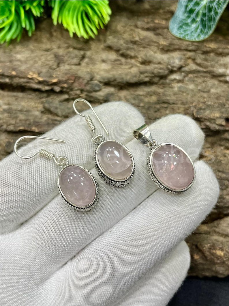May include: A jewelry set featuring a pair of earrings and a pendant. Each piece has a light pink, oval-shaped stone set in a silver-colored metal frame. The earrings have a hook design, and the pendant has a loop for a chain.