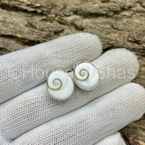 Peut inclure: Une paire de boucles d'oreilles blanches et argentées. Chaque boucle d'oreille présente un motif ovale blanc avec un motif en spirale au centre. Les boucles d'oreilles sont serties dans un cadre en métal argenté.