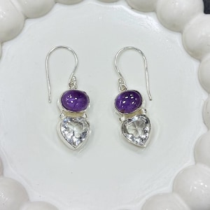 Può includere: Orecchini in argento con una gemma ovale viola e una gemma trasparente a forma di cuore. Gli orecchini hanno un design a gancio e sono presentati su un piatto bianco con bordo smerlato.