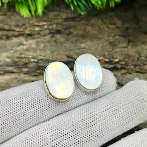 以下が含まれることがあります： 楕円形のスタッドピアス。各イヤリングは、銀色の金属フレームにセットされた、きらめく虹色の真珠母貝の中心部を備えています。イヤリングは、白くテクスチャ加工された表面に表示されています。