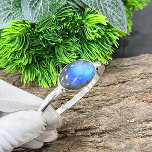 May include: A silver bracelet featuring an oval labradorite stone with blue and iridescent flashes. The bracelet is held against a backdrop of green foliage and a textured wooden surface.