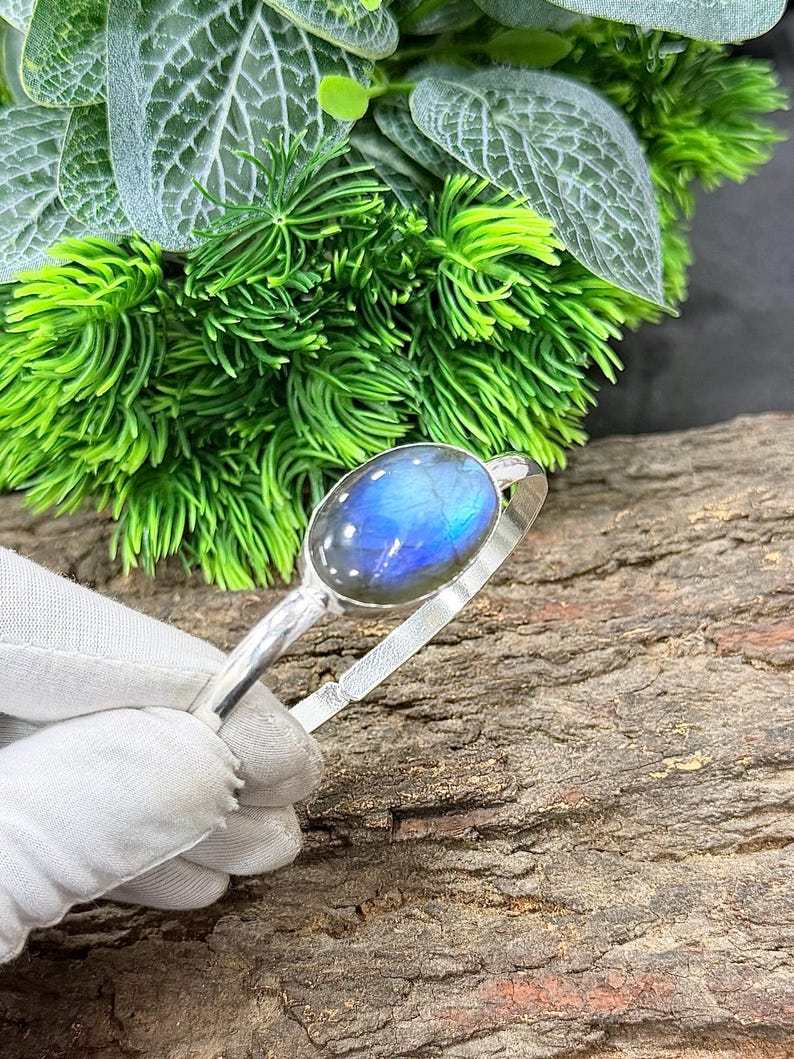 May include: A silver bracelet featuring an oval labradorite stone with blue and iridescent flashes. The bracelet is held against a backdrop of green foliage and a textured wooden surface.
