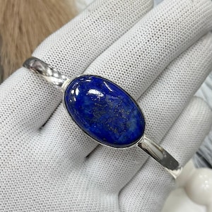 May include: A silver cuff bracelet featuring a large, oval lapis lazuli stone. The deep blue stone has gold flecks and is set in a polished silver band. The bracelet is shown on a white glove.