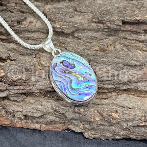 Peut inclure: Collier avec pendentif ovale et chaîne argentée. Le pendentif présente une coquille d'ormeau irisée aux nuances de bleu, violet et vert. La coquille est sertie dans un cadre argenté. Le collier est présenté sur une surface en bois brun texturé.