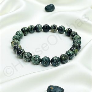 May include: A beaded bracelet featuring dark green and black speckled beads. The bracelet is made of round, polished stones and is displayed on a white satin surface. Two loose stones are also present.