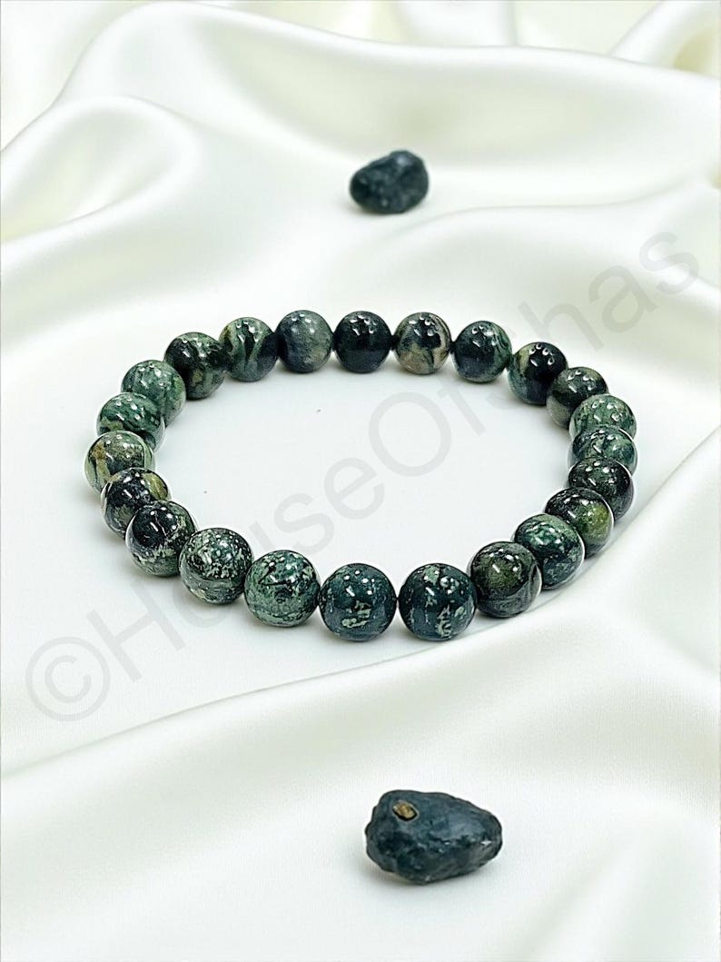 May include: A beaded bracelet featuring dark green and black speckled beads. The bracelet is made of round, polished stones and is displayed on a white satin surface. Two loose stones are also present.
