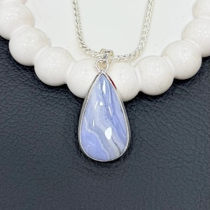 May include: A silver necklace featuring a teardrop-shaped pendant. The pendant has a light blue stone with white and gray marbling, set in a silver bezel. The chain is a delicate, woven design. The necklace is displayed on a white, beaded surface.