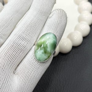 May include: A silver ring featuring an oval-shaped, light green gemstone with white marbling. The ring is set on a silver band and is displayed on a white glove. The gemstone has a polished, glossy finish.