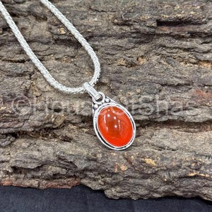 Könnte beinhalten: Eine silberne Anhängerkette mit einem ovalen, orangerotfarbenen Edelstein. Der Anhänger ist in einer silbernen Lünette gefasst und hängt an einer zarten Silberkette. Die Halskette wird auf einer strukturierten, braunen Holzoberfläche präsentiert.