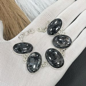 Pode incluir: Uma pulseira de prata com seis pedras ovais cinza escuro com um padrão salpicado. As pedras são conectadas por uma delicada corrente de prata. A pulseira é exibida em uma superfície branca.