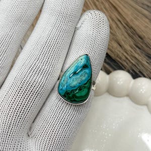 May include: A teardrop-shaped ring featuring a vibrant turquoise and green stone set in a silver band. The stone's surface displays a mix of blue and green hues, creating a unique pattern. The ring is shown on a white glove.