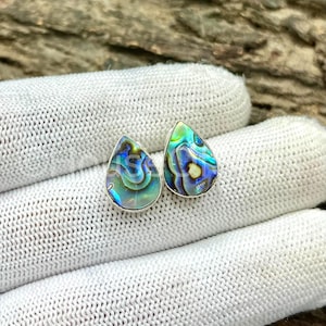 May include: A pair of teardrop-shaped abalone shell earrings. The earrings have a silver-colored border and display iridescent blues, greens, and yellows. The earrings are displayed on a white, textured surface.