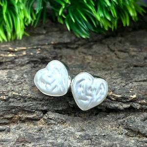 May include: A pair of heart-shaped earrings with a pearlescent white finish. The earrings are set against a textured, dark brown background, with green foliage in the upper portion of the image.