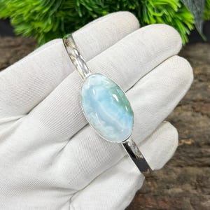 May include: A silver bracelet featuring a large, oval-shaped larimar stone. The stone displays a mix of light blue and white hues, with a marbled appearance. The bracelet is set against a white background, highlighting the jewelry's design and color.
