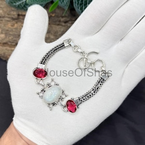 Peut inclure: Un bracelet en argent orné d'une pierre précieuse blanche ovale au centre, flanquée de deux pierres rouges ovales. Le bracelet a un design en forme de chaîne. Le bracelet est un bijou.