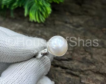May include: A silver ring featuring a large, luminous white pearl. The pearl is round and set in a simple, elegant band. The ring is held by a gloved hand against a backdrop of wood and green foliage.