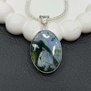 May include: Oval pendant necklace with a polished moss agate stone, displaying green and white patterns. The stone is set in a silver-coloured metal frame and hangs from a silver chain. The necklace is on a white, textured surface.