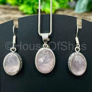 May include: A jewelry set featuring a rose quartz pendant necklace and matching earrings. The oval-shaped stones are set in silver-toned metal with a beaded border. The earrings have a hook closure. The necklace has a delicate chain.