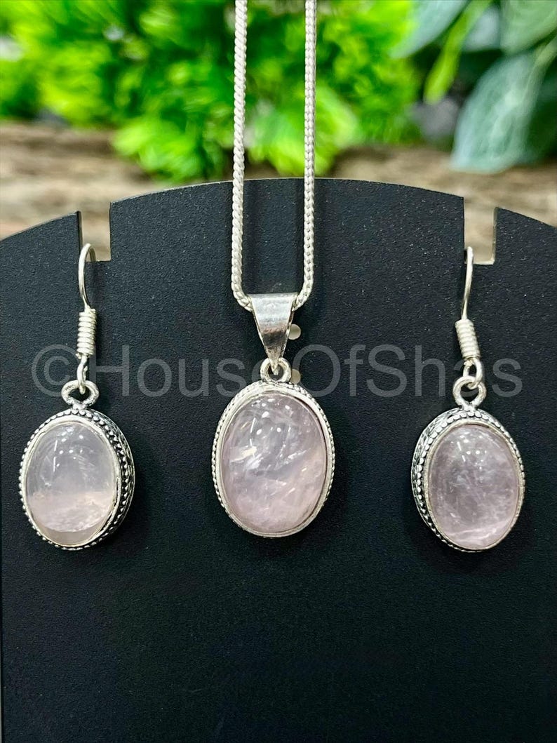 May include: A jewelry set featuring a rose quartz pendant necklace and matching earrings. The oval-shaped stones are set in silver-toned metal with a beaded border. The earrings have a hook closure. The necklace has a delicate chain.