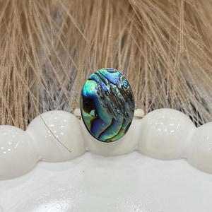 May include: Oval-shaped ring featuring an iridescent abalone shell set in a silver band. The shell displays a mix of blue, green, and purple hues. The ring is displayed on a white, scalloped surface, with a soft, feathery background.