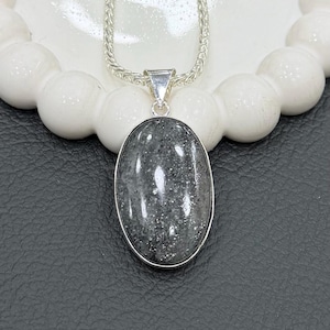 Può includere: Una collana d'argento con un pendente ovale. Il pendente presenta una pietra grigio scuro con un motivo maculato, incastonata in una lunetta d'argento. La catena è di stile fine e intrecciato. La collana è esposta su una superficie bianca strutturata.