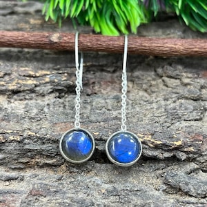 May include: Silver drop earrings with round, iridescent blue labradorite stones. The stones are set in silver bezels and hang from delicate silver chains. The earrings are displayed on a textured, brown wooden surface.