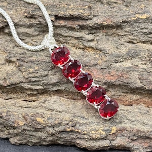 May include: A silver necklace featuring a series of five oval-cut, deep red gemstones. The gemstones are set in a silver pendant that hangs from a delicate silver chain. The necklace is displayed against a textured, brown background.