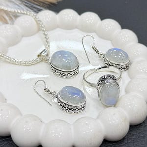以下が含まれることがあります： ネックレス、イヤリング、指輪のジュエリーセット。すべてオーバル型のイリデッセントムーンストーンカボションが装飾的なシルバーメタルにセットされています。セットは白い装飾トレイに展示されています。