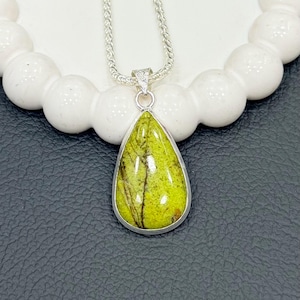 May include: A silver pendant necklace featuring a pear-shaped, lime green gemstone with brown veining. The stone is set in a silver bezel and hangs from a delicate silver chain. The necklace is displayed on a white, textured surface.