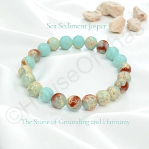 May include: A beaded bracelet featuring Sea Sediment Jasper stones. The bracelet alternates between light blue and speckled beige and red beads. The text "Sea Sediment Jasper" and "The Stone of Grounding and Harmony" are visible.