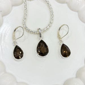 May include: A silver necklace and earring set. The necklace has a chain and a teardrop pendant with a dark brown gemstone. The earrings also have teardrop-shaped dark brown gemstones. The set is displayed on a white, scalloped plate.