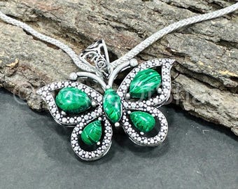 Pode incluir: Um colar com pingente de borboleta prateada com pedras de malaquite verde. O design da borboleta apresenta asas detalhadas e um corpo central, com as pedras verdes adicionando um toque de cor. O colar é exibido em uma superfície de madeira texturizada.