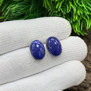 May include: Two oval-shaped, deep blue lapis lazuli stud earrings with silver settings. The earrings are displayed on a white knitted surface, with green foliage in the background.