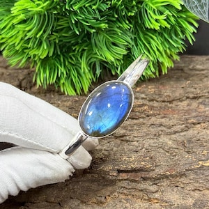 May include: A silver bracelet featuring an oval labradorite gemstone with blue and iridescent flashes. The gemstone is set in a polished silver band. The bracelet is displayed against a natural wood background with green foliage.