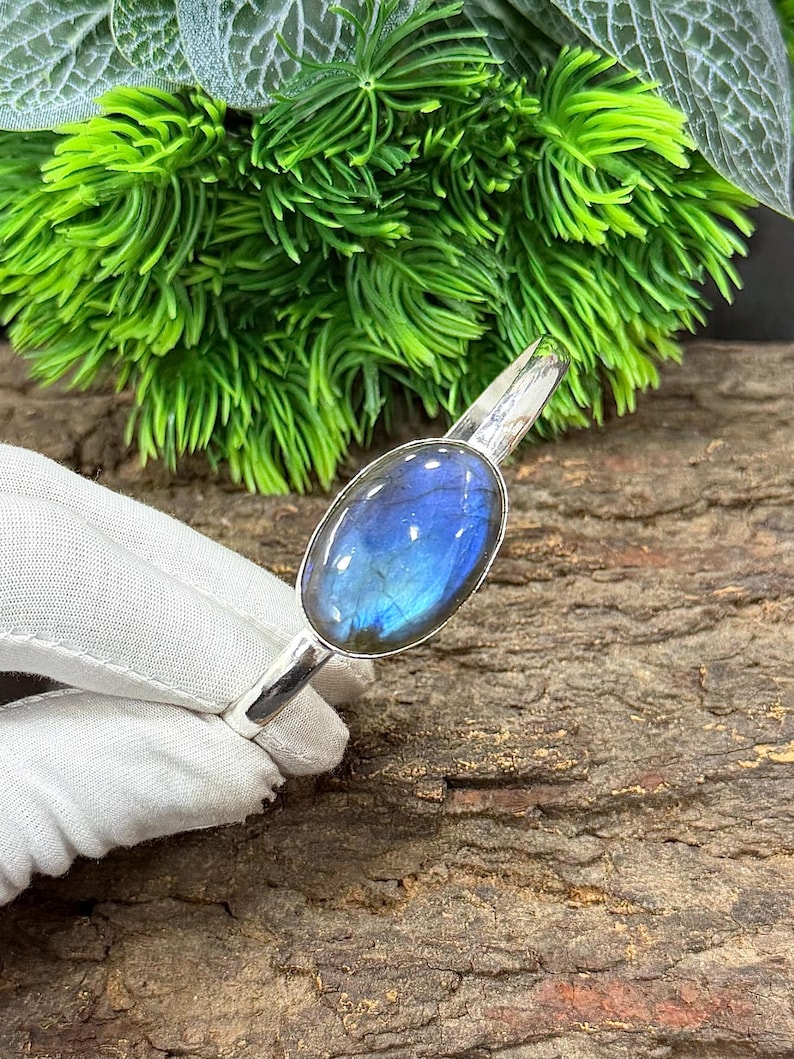 May include: A silver bracelet featuring an oval labradorite gemstone with blue and iridescent flashes. The gemstone is set in a polished silver band. The bracelet is displayed against a natural wood background with green foliage.