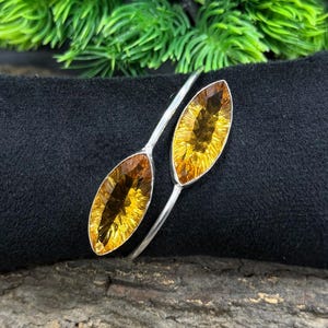 May include: A silver bracelet with two oval-cut citrine gemstones. The gemstones are a warm yellow-orange hue, set in a minimalist design. The bracelet is displayed on a black surface, with green foliage in the background.