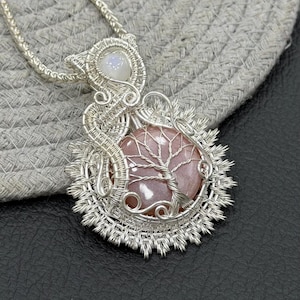 May include: A silver wire-wrapped pendant necklace featuring a round, peach-colored stone with a wire tree design on its surface. The pendant is accented with a small, iridescent white cabochon at the top and intricate, radiating silver wirework.