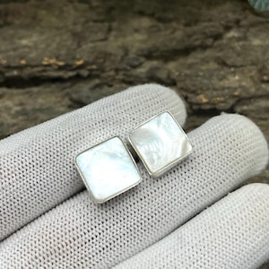 May include: A pair of square earrings with a silver border. Each earring features a pearlescent, white, square centre. The earrings are displayed on a white, knitted surface.