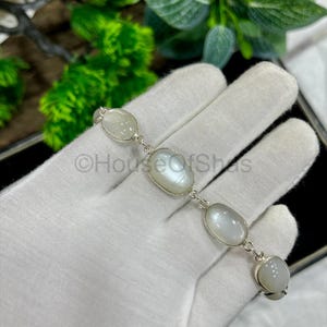 Puede incluir: Un brazalete de plata con cabujones ovalados de piedra lunar iridiscente. El brazalete está compuesto por una serie de piedras enlazadas, cada una engastada en un bisel de plata. El brazalete se exhibe sobre una superficie blanca.