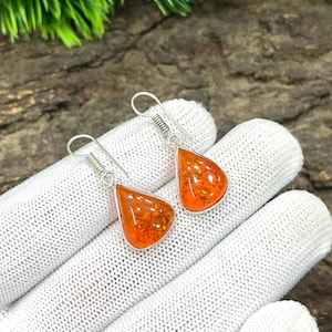 May include: A pair of silver dangle earrings featuring teardrop-shaped orange amber stones. The amber has a speckled, textured appearance. The earrings are displayed on a white surface, with a natural wood background.