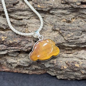 May include: A silver chain necklace with a cloud-shaped pendant. The pendant is a polished, golden-brown gemstone. The necklace is displayed on a textured, brown surface.